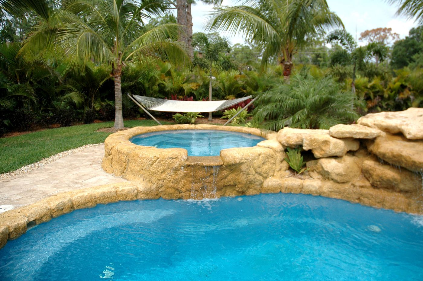 Freeform Laguna fiberglass pool and spa with tropical rock waterfall landscaping and a white backyard hammock.