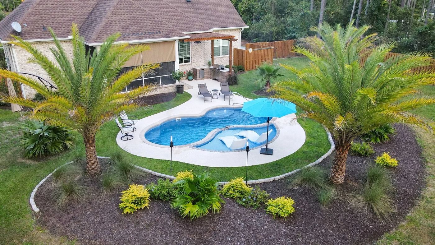 Freeform fiberglass pool surrounded by palm trees, featuring a tanning ledge and a single blue umbrella.